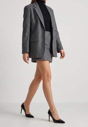 Kvinde gående iført en grå blazer, matchende grå nederdel, sort rullekrave og sorte højhælede sko mod en ensfarvet baggrund.