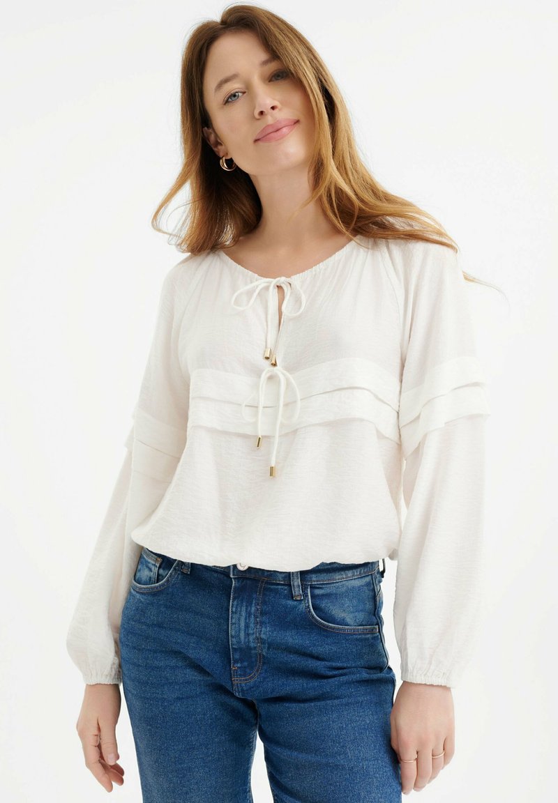 Blusa blanca de manga larga con cuello redondo, detalle frontal con cordón y paneles horizontales en capas. Combinada con jeans de mezclilla azules.