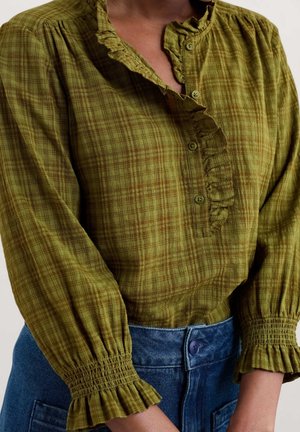 Groene geruite blouse met lange mouwen, gerimpelde kraag en manchetten, ingestopt in een hooggetailleerde blauwe spijkerbroek.