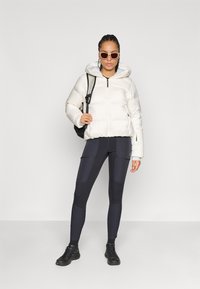 Biela puffrová bunda s kapucňou, čierne legíny s detailmi vrecka, čierny batoh a čierne športové topánky. Príslušenstvo zahŕňa slnečné okuliare.