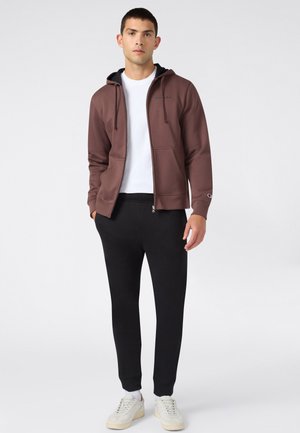 Jonge man staat met een bruine hoodie met rits, wit T-shirt, zwarte joggingbroek en witte sneakers op een effen achtergrond.