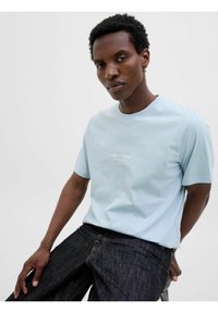 Light blue short-sleeve t-shirt with a crew neck and white "Originals Studio" logo on the chest, paired with dark denim jeans.