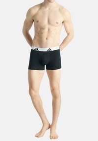 Svarta boxerkalsonger i elastiskt tyg, med ett vitt elastiskt midjeband som har svarta Adidas-logotypdetaljer. Längd till mitten av låret.