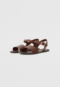 Brune lær sandaler med flad sål, der har brede remme over foden og en justerbar spændeankelrem. Glat tekstur.