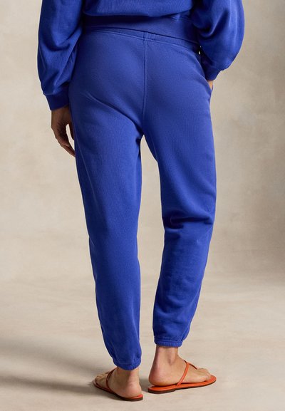 Osoba w jaskrawo niebieskich spodniach dresowych i pasującej bluzie, stojąca boso w pomarańczowych płaskich sandałach na neutralnym tle.