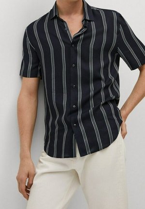 Homme portant une chemise noire à manches courtes avec des rayures blanches verticales et un pantalon écru, posant avec une main sur la hanche.