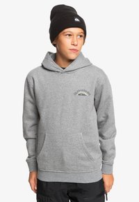 Sudadera con capucha gris, con un bolsillo frontal, logo bordado y puños de canalé. Lleva un gorro negro. El tejido parece suave y texturizado.