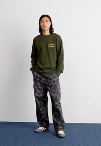 Sweater canelado verde-oliva com texto amarelo, combinado com calças cargo cinza camuflado. Inclui um corte descontraído e bolsos laterais. Ténis brancos de desporto.