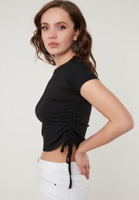 Zwarte geribbelde top met korte mouwen, voorzien van zijruche en verstelbare strikken. Draag met een witte jeans, die een aansluitende silhouet toont.