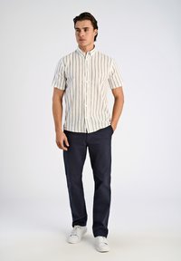Kurzarm-Hemd mit Knöpfen in Creme mit grauen vertikalen Streifen, kombiniert mit navyblauen Hosen und weißen Sneakers, vor einem schlichten Hintergrund stehend.