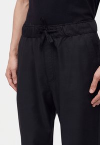 Homme portant un pantalon noir avec une taille élastique et un cordon noué, les mains détendues sur les côtés, assorti à une chemise noire.