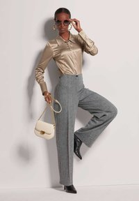 Beige satijnen overhemd met knopen, grijze wijde broek, zwarte puntige hakken en een crèmekleurige handtas met een kettingband. Circulaire zonnebril.