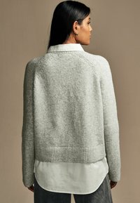 Grå strikket sweater med en boxy pasform, ribbet kant og strukturerede detaljer. Bæres over en hvid skjorte med krave og et lagdelt udseende.