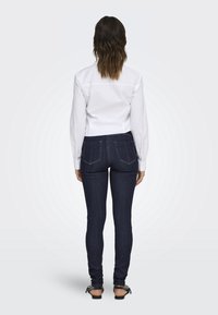 Camisa blanca de manga larga y jeans ajustados de mezclilla oscura, con un diseño clásico de 5 bolsillos, costuras en contraste y un ligero brillo.