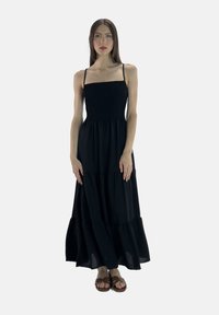 Sort maxi-kjole med spaghetti-stropper, smocket bodice, tiered nederdel og en afslappet pasform. Iført med brune sandaler. Ensfarvet baggrund.