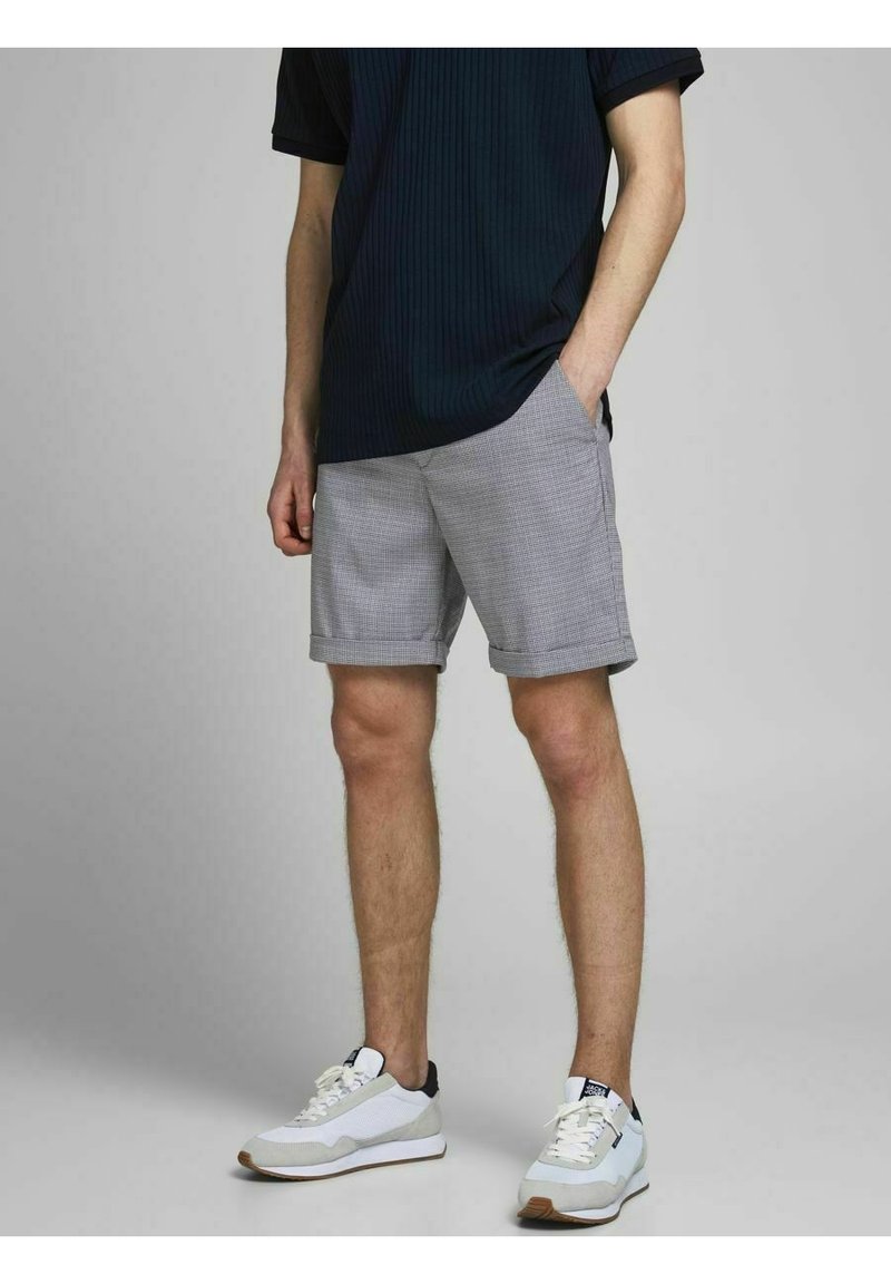 Homme portant un short gris à carreaux, un t-shirt noir et des baskets blanches, debout avec une main dans la poche devant un fond uni.