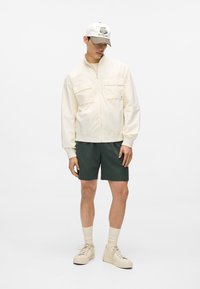 Giacca bomber color crema chiaro con due tasche sul petto, abbinata a pantaloncini verde scuro e sneakers beige. Il cappellino presenta un ricamo.