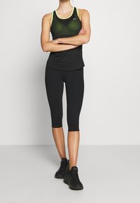 Canotta sportiva nera con design in rete e dettagli giallo neon, abbinata a leggings neri di media lunghezza e scarpe da ginnastica nere.
