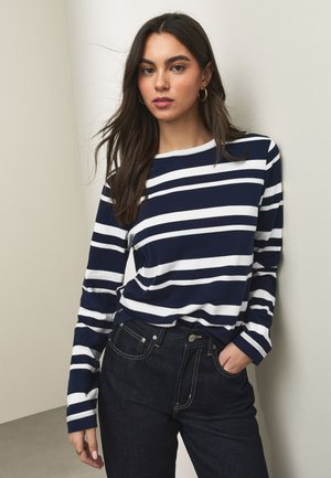 Žena s dlhými tmavými vlasmi, ktorá má na sebe zlaté kruhové náušnice, tmavomodro-biele pruhované tričko s dlhým rukávom a tmavomodré džínsy, pričom jednu ruku má v vrecku.