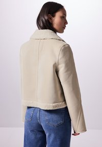 Avskuren, krämfärgad läderjacka med en shearlingkrage och manschetter, som har en slät yta och rena linjer. Buren med blå jeans.