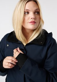 Marineblauwe waterdichte jas met een zwart fleece gevoerd kraag, ritsluiting en aansluitende manchetten. Bevat metalen drukknopen en een gladde textuur.