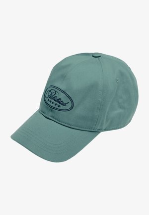 Jeune homme portant une casquette verte avec le texte "Chabilited" et des étoiles, chemise rayée à boutons par-dessus un t-shirt blanc, fond neutre.