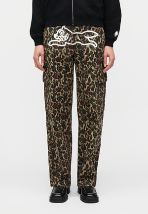 Kargo hlače s kamuflažnim vzorcem in veliko risano podobo psa z belim robom na sprednji strani, nošene s črnimi čevlji in črno jakno.