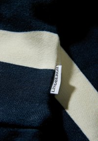 Dzianina w paski w kolorach granatowym i kremowym o miękkiej fakturze. Zawiera białą metkę z napisem "LINDBERGH" wszytą w szew.