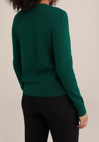 Grön stickad tröja med ett texturerat mönster, rund halsringning och ribbad nederkant och ärmslut, kombinerad med svarta figurnära byxor.