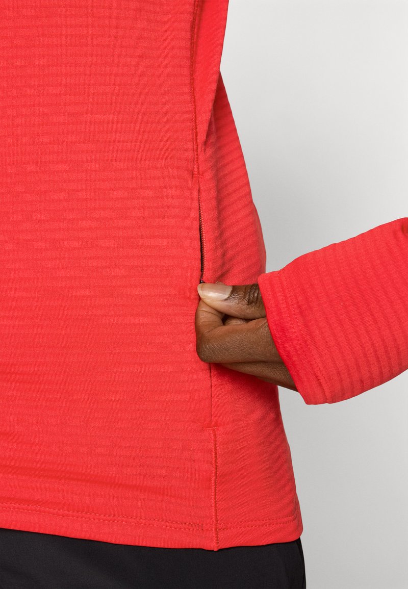 Czerwona koszula z długim rękawem o teksturowanym wzorze, z boczną kieszenią i otworami na kciuki przy mankietach.