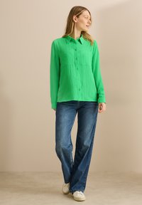Camicia verde a maniche lunghe, realizzata in tessuto testurizzato, con due tasche frontali, abbinata a jeans blu e sneakers bianche.
