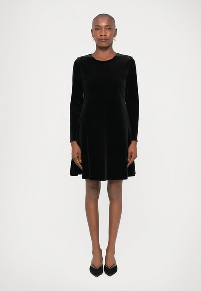 Robe noire en velours à manches longues avec un décolleté arrondi et une jupe évasée. Associée à des escarpins noirs à bouts pointus, sur fond blanc uni.