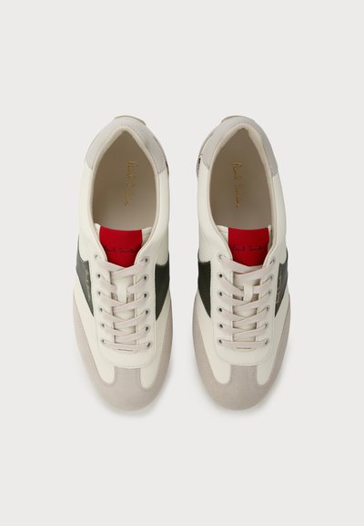 Paire de baskets blanches et grises avec des bandes latérales noires et des étiquettes rouges sur la languette, vues de dessus sur un fond clair.