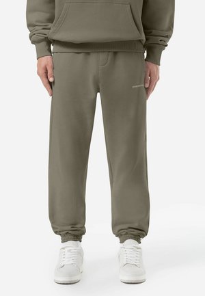 Olivengrønne sweats med elastisk talje og manchetter, sidelommer og et lille logo nær hoften. Stoffet ser blødt og glat ud.