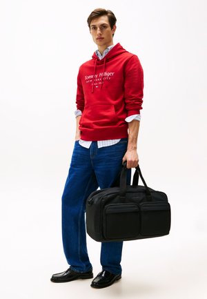 Svart nylon gymbag med doble frontlommer, solide håndtak og logo-detaler. Modellen har på seg en rød hettegenser, blå jeans og svarte sko.