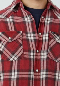 Chemise à carreaux rouge et blanche avec deux poches poitrine, boutons pression en nacre, et une texture en coton. Présente des motifs à carreaux contrastés foncés et clairs.