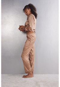 Beige satin lounge-outfit set met een langetoff top met ronde hals en broek met een elastische tailleband en cuffs aan de onderkant, tegen een grijze achtergrond.