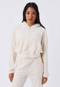 Sudadera cremosa con cremallera, hecha de un tejido suave, que cuenta con una capucha con cordones y puños elásticos. Pequeño detalle de logo metálico en el pecho.