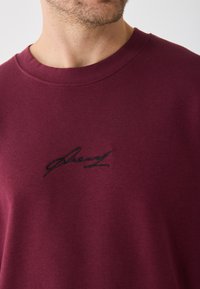 Sweat-shirt bordeaux au toucher doux, doté d'un col rond et d'un détail de signature brodé noir à l'avant.
