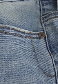 Mörkblå denimtyg med en synlig ficka, bruna sömmar och en metallknapp märkt "INDIGO BLUE JEANS." Texturerat vävmönster.