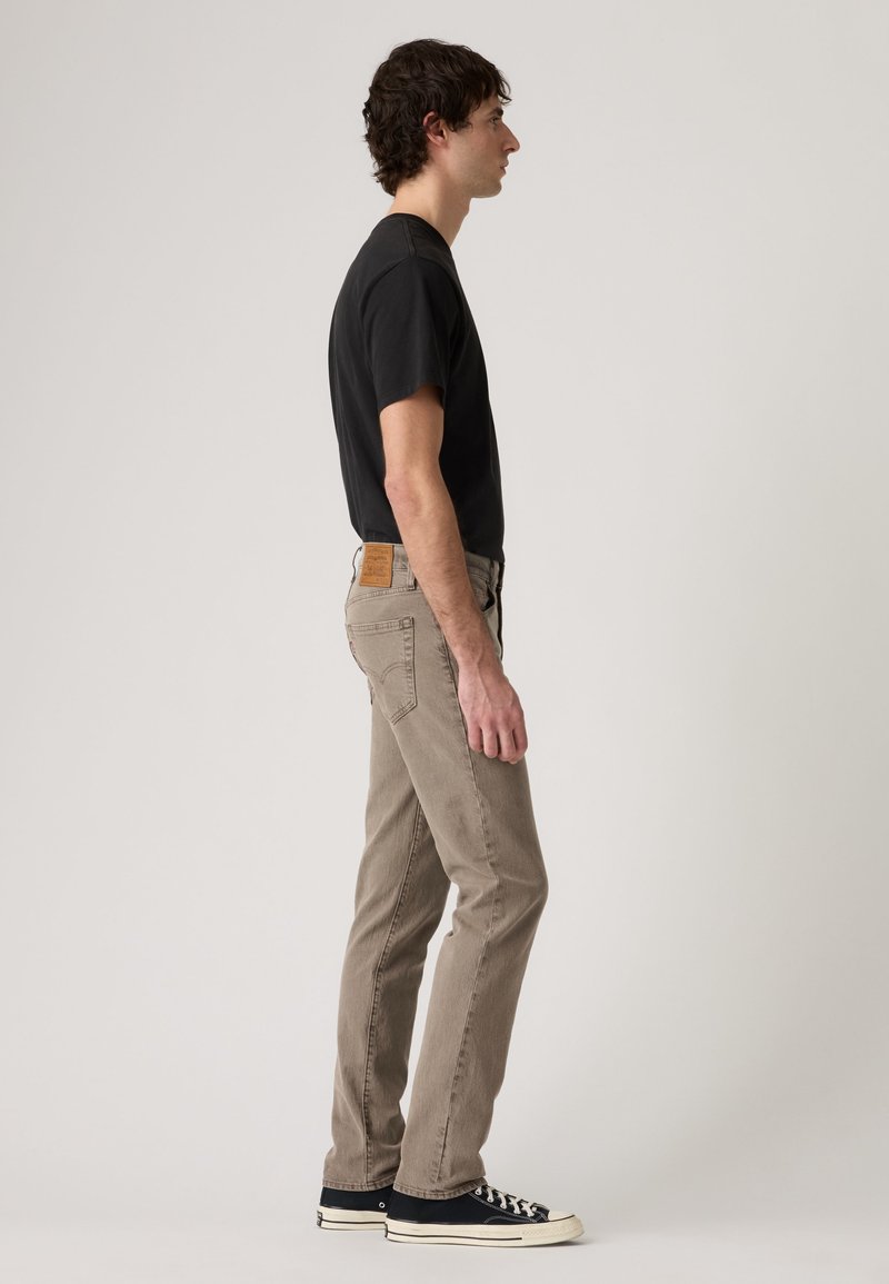 Uomo in piedi di profilo che indossa una t-shirt nera, jeans beige e scarpe sportive basse nere su sfondo chiaro uniforme.