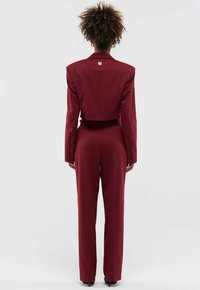 Vrouw met krullend haar draagt een op maat gemaakte bordeauxrode cropped blazer en bijpassende hoog-getailleerde broek, staand met de rug naar de camera op een effen achtergrond.