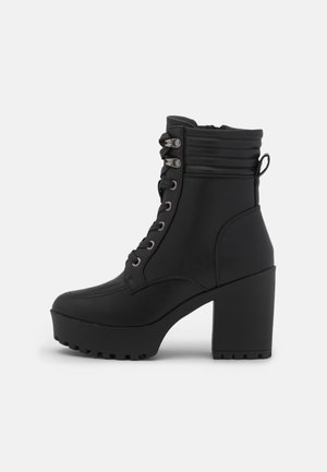 Bottine noire à talon épais avec devant lacé, col matelassé, fermeture éclair latérale et languette sur fond blanc.