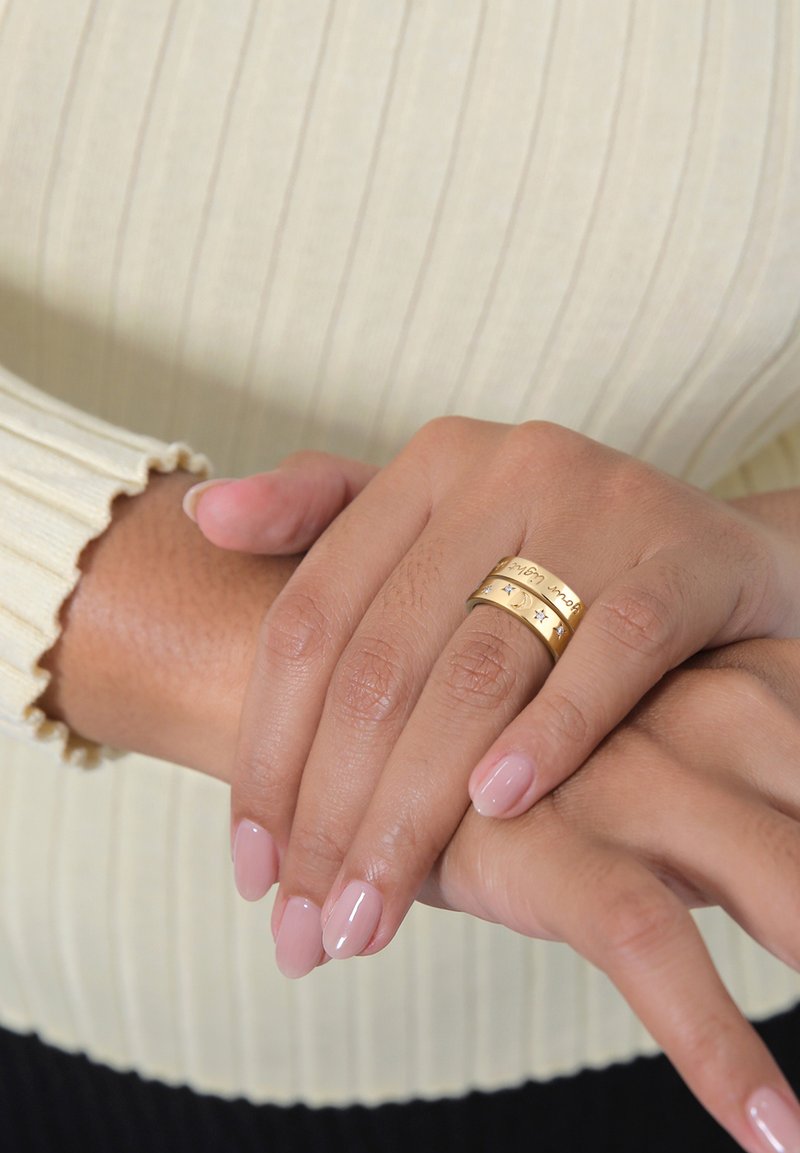 Anello color oro con tre bande sovrapposte, caratterizzato da incisioni e piccoli accenti a forma di stella. Le mani hanno una finitura naturale con unghie di un leggero colore rosa.