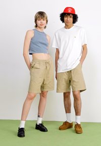 Two individuals pose side by side. One wears a blue cropped tank top and beige shorts; the other wears a white T-shirt and beige shorts.