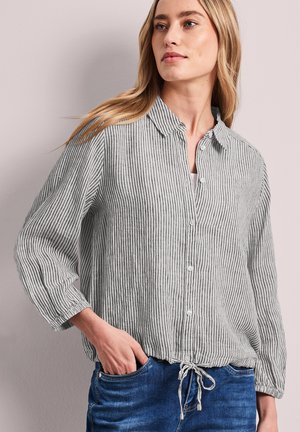 Femme aux longs cheveux blonds portant une chemise à rayures verticales grises et blanches boutonnée et un jean bleu, une main dans la poche, regardant sur le côté.