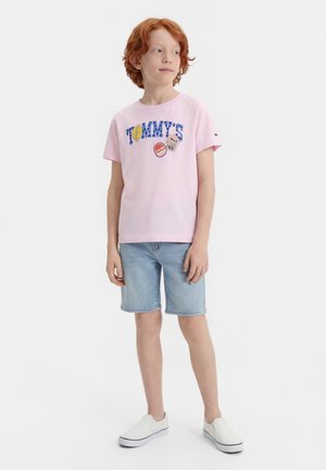 Ragazzo dai capelli rossi in piedi, indossa una maglietta Tommy rosa chiaro con toppe, pantaloncini di jeans azzurri e scarpe slip-on bianche, guarda di lato.