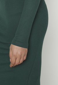 Personne portant une robe ajustée à manches longues côtelée de couleur vert foncé, la main gauche détendue le long du corps.