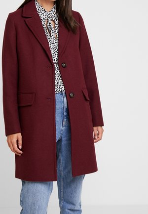 Kobieta ubrana w bordowy wełniany płaszcz na wzorzystej czarno-białej bluzce i jasnoniebieskich dżinsach, stojąca na jednolitym tle.