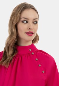 Lyserød bluse med høj krave og plisseringer, dekoreret med sølvfarvede blomsterformede knapper langs skulderen. Glat stofstruktur.